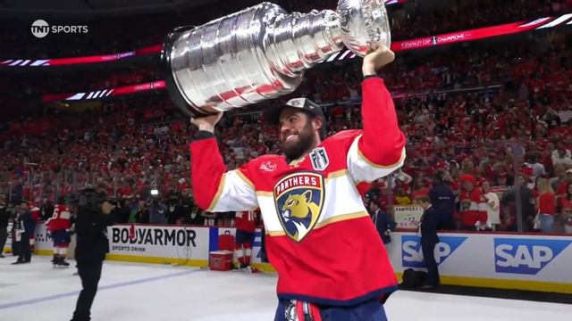 A bearded Florida Panthers player in a red jersey and black championship cap joyfully lifts the Stanley Cup high over his head while skating across the ice. The shiny silver trophy glows under arena lights, and the stands are packed with fans dressed in red, many waving and cheering. The boards display ads including “BodyArmor” and “SAP,” and a photographer captures the scene nearby.