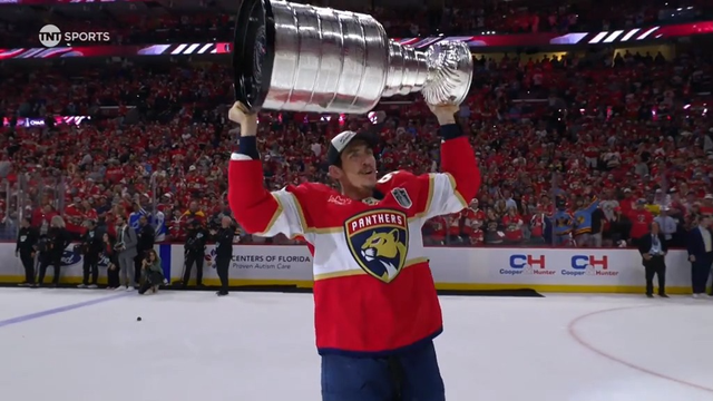 A Florida Panthers player with a red beard stands proudly at center ice, arms raised as he holds the Stanley Cup above his head. The underside of the trophy is visible, featuring the NHL logo. Behind him, the entire Panthers team claps and celebrates, all wearing red jerseys and white caps marked “Champions.” The crowd in the packed arena roars in the background.