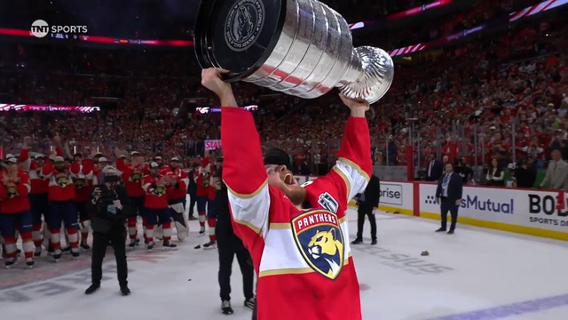 A smiling Florida Panthers player in a white championship cap lifts the Stanley Cup overhead with both arms. He wears a red jersey and stands among teammates on the ice. Behind him, other players and photographers are visible as the championship celebration continues. His joyful expression captures the thrill of victory on hockey’s biggest stage.