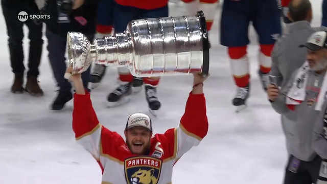 Another Florida Panthers player, beaming with pride, holds the Stanley Cup high above his head. The crowd behind him is a sea of red, packed into the arena and cheering loudly. He wears the team’s red and gold jersey with the panther logo prominently displayed. The boards behind show a “CH Cooper Hunter” ad as photographers snap pictures of the moment.