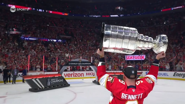 Florida Panthers forward Sam Bennett stands proudly on the ice holding the Stanley Cup high above his head with both hands. He wears a red jersey with his name and number (9) visible on the back. Behind him, a packed arena of cheering fans is lit by red lights, and the Stanley Cup Champions stage and signage are seen in the distance. The scene captures jubilation and triumph.