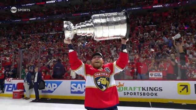 A bearded Florida Panthers player beams with pride as he hoists the Stanley Cup above his head in front of a sea of cheering fans. He wears a red jersey with the team’s logo and a white cap labeled “Champions.” The boards behind him are lined with ads from SAP and DraftKings, and the crowd is electric with excitement.