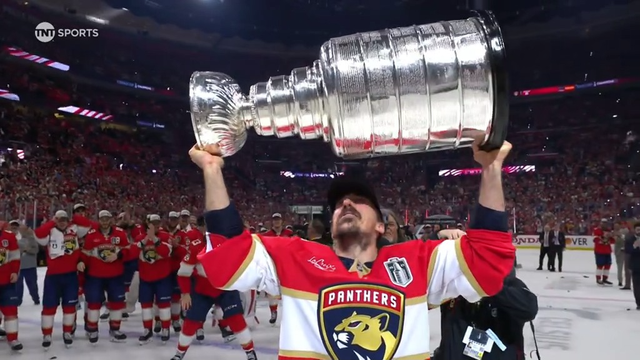 A Florida Panthers player in a red jersey raises the Stanley Cup above his head while looking up at it with admiration. His teammates, also in red jerseys and white championship caps, cheer and applaud in the background. The arena is filled with fans celebrating the Panthers’ historic championship win.