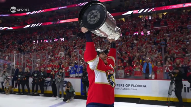 Another Florida Panthers player holds the Stanley Cup aloft with both arms while smiling joyfully. He stands in front of a section packed with fans dressed in red, many clapping and capturing the moment. The underside of the Cup with the NHL seal is visible. Photographers along the glass document the scene, and the boards read “Centers of Florida.”