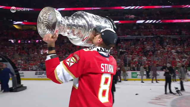 Florida Panthers player Nico Sturm, wearing a red jersey with the number 8 and his name on the back, tilts the Stanley Cup back to kiss it in celebration. He stands on the ice facing a packed arena full of fans dressed in red. The iconic silver trophy gleams under the arena lights. It’s a moment of pride, emotion, and tradition.