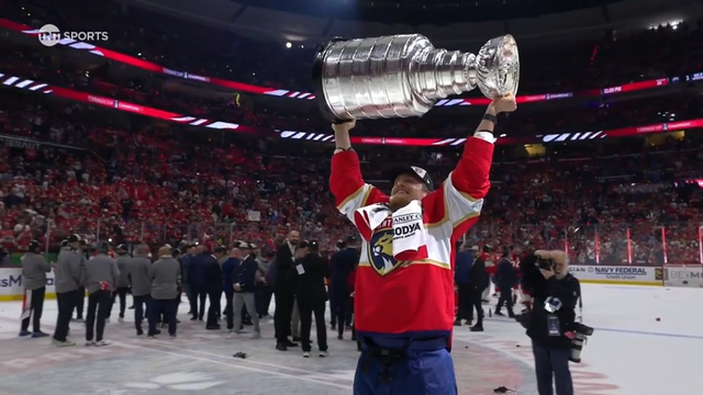 A Florida Panthers player celebrates by hoisting the Stanley Cup high above his head. Draped in a white towel labeled “Stanley Cup Champions,” he grins with joy as photographers and teammates gather on the ice. Fans cheer in the packed arena behind him. Confetti and gear are scattered across the ice as the celebration continues.