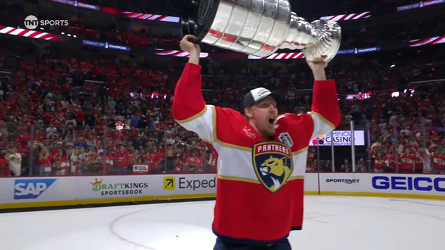A Florida Panthers player roars with excitement as he lifts the Stanley Cup over his head with both arms. He wears the team’s red jersey and a white “Champions” cap, with a beaming smile and clenched fists. Behind him, a vibrant crowd of fans and a wall of advertisements, including SAP and DraftKings, fill the background of the electric scene.