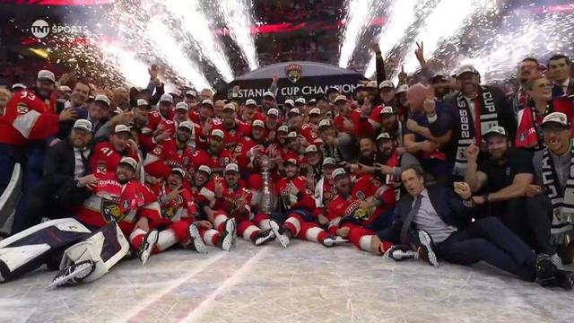The entire Florida Panthers team and staff gather tightly at center ice for a celebratory group photo after winning the 2025 Stanley Cup. All are wearing red jerseys and white championship caps, many with arms raised, smiling and cheering. The massive silver Stanley Cup trophy is placed prominently in the center. Behind them, golden fireworks erupt skyward, and a black backdrop reads “STANLEY CUP CHAMPIONS.” The ice is littered with gear and celebration remnants, capturing the peak moment of victory and unity.