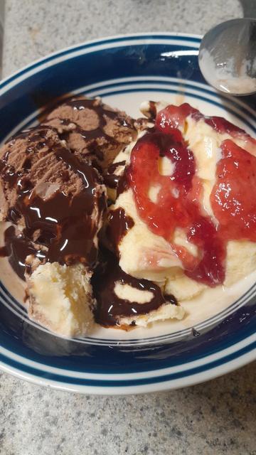 A bowl with chocolate ice cream and chocolate syrup on one side and vanilla ice cream with strawberry preserves on the other and a spoon hanging over the top