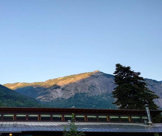 Berge, auf dem rechten Gipfel ist ein Gebäude erkennbar.
Im Vordergrund steht rechts ein Baum. Am unteren Bildrand ist ein Dach. Es ist blauer Himmel. 