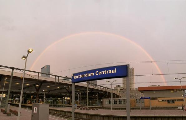 Dubbele regenboog in een grijze hemel, op de voorgrond stationsbord Rotterdam Centraal.