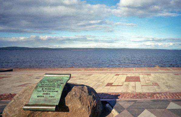 In the foreground is the embankment, laid with granite slabs of warm shades. On it stands a large stone, to which a copper, green plate in the form of a scroll is attached. Text is engraved on the plate. In the background is a strip of dark blue water of Lake Onega. In the background is visible the other shore. Blue sky with clouds.