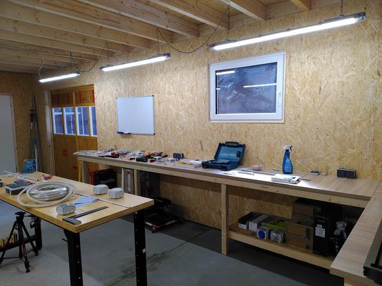 New shop showing window and whiteboard above a wood workbench. In the far left side of the image is a large sliding door with 4 windows. In the middle of the room is a metal workbench with a wooden top.