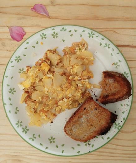 A plate of scrambled eggs mushrooms and buttered toast, on a pale wood surface, seen from above. The playe is white with a green leaf pattern, the toast is sourdough and slightly burnt.