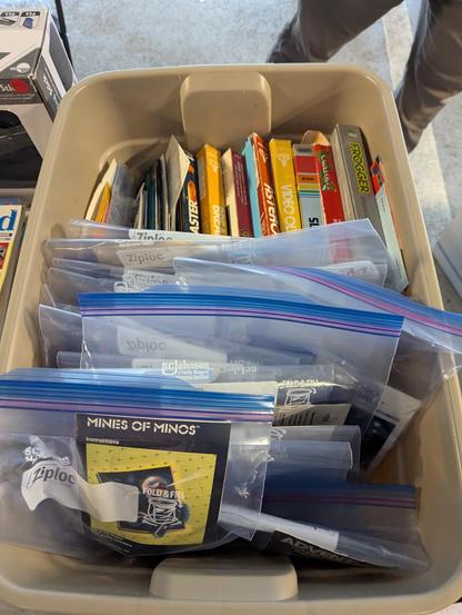 A photograph of a bin of software for Atari systems.  Some of it is in its original packaging, some is in individual Ziploc bags.  Foremost in the photo is a bag with "Mines of Minos."