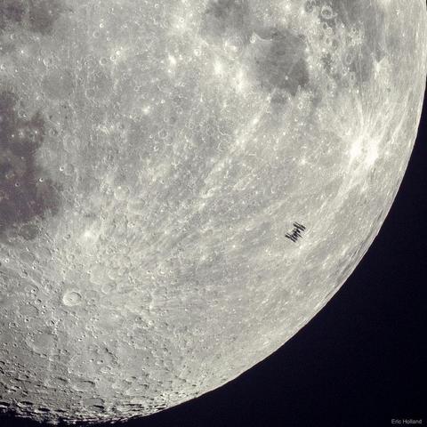 A high-resolution image of the Moon showing the silhouette of the International Space Station (ISS) transiting in front of it. The ISS appears as a tiny, dark, sharply outlined shape with visible solar panels and structural elements, frozen mid-transit. The Moon is partially lit in a gibbous phase, showing detailed surface features including the bright crater Tycho in the lower left, along with rough highland terrain and darker maria regions.