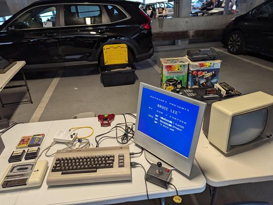 A photograph of a vendor table at the MIT Flea on 2025.06.15, with a complete Commodore 64 system arrayed and running, together with cartridges, a joystick, and a tape deck.  It is connected to a modern flat panel display that is showing the menu screen for "Bruce Lee" by Datasoft.