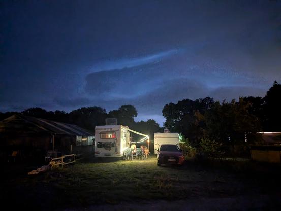 Das Bild zeigt eine nächtliche Szene auf einem Campingplatz. Im Vordergrund sieht man zwei Wohnmobile, die mit Lichtern geschmückt sind. Vor einem der Wohnmobile sitzen einige Personen an einem kleinen Tisch und scheinen sich zu unterhalten oder eine Mahlzeit einzunehmen. Ein Auto ist neben den Wohnmobilen geparkt. Im Hintergrund sind einige Bäume und ein Gebäude zu erkennen. Der Himmel ist dunkel mit einigen Wolken, die von einer entfernten Lichtquelle beleuchtet werden, möglicherweise von einer Stadt oder einem Gewitter. Die Atmosphäre wirkt ruhig und gemütlich.