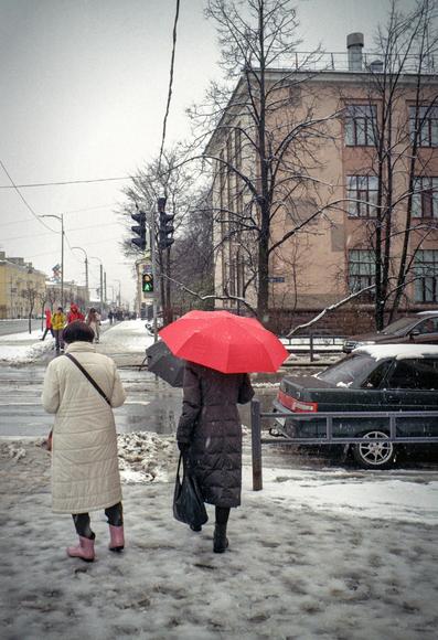 In the photo, the women are standing at an intersection in the city. One of them is holding a red umbrella. It's snowing wet.
