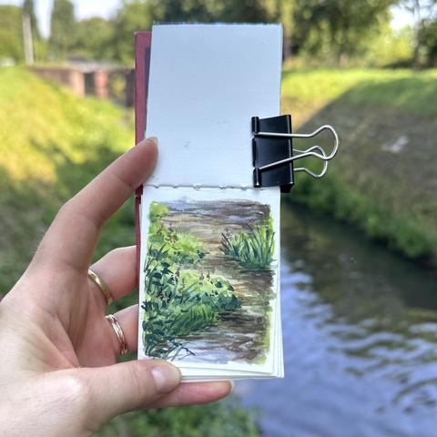 A photo of my hand holding up a tiny sketchbook in which I painted part of the overgrown canal that can be seen blurrily in the background.