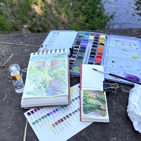 A photo of two sketchbooks of different sizes and some art supplies placed on a cement outcrop next to a little canal that a small bit can be seen of on the top right. The left sketchbook shows my friend’s rendition of the view, the right one mine.