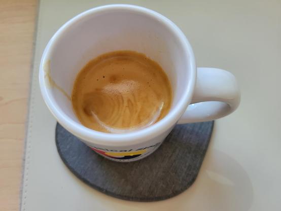 A white, 3 ounce, ceramic espresso (demitasse) cup with a shot of espresso, on a gray diatomaceous-earth coaster on an off-white desk mat. The blue text on the cup reads "Cyber-Physical Systems Research Center" and wraps in a circle around the logo, which looks like a loosely interlaced, rounded triquetra with thick knots in solid red, yellow, and blue (each its own color). 