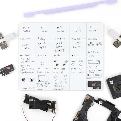 A white magnetic project mat is organized with handwritten labels and neatly sorted screws from a disassembled device. Each section of the grid corresponds to a specific component—like the battery, speakers, fan, and hinge—along with the screw type and quantity used. Surrounding the mat are various internal laptop parts, including speakers, a purple bezel, a fan, and circuit boards.