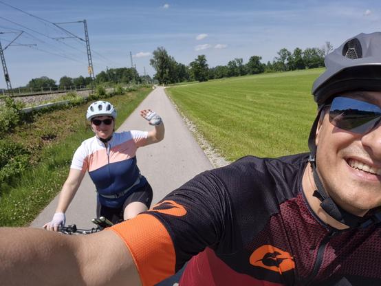 A man and woman are seen riding bikes on a road in this outdoor scene. The image features a man and woman, both wearing helmets and sunglasses, smiling as they pedal along. The background consists of grass and trees under a clear sky.
