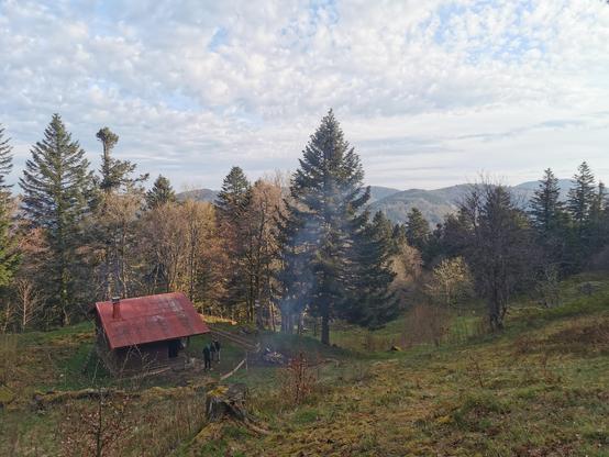 Une cabane isolée dans la montagne vosgienne, vue à distance. Devant la cabane s'élève la maigre fumée dun feu de camp.