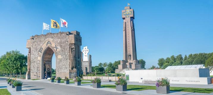 Panoramic view of the Yser Tower (IJzertoren) in Diksmuide, Belgium, on a bright, sunny day with clear blue skies. The tall, cross-shaped brick tower stands prominently in the background, symbolizing peace and Flemish remembrance of World War I. Large yellow letters “AVV-VVK” (a Flemish slogan meaning "Everything For Flanders, Flanders For Christ") are displayed near the top of the tower. In front of it stands the remains of the original, white-coloured Yser Tower, and the "Gate of Peace", made of dark stone with a large arched opening and the word “PAX” (Latin for “peace”) above it. Three flags fly above the gate: one with the Flemish lion, one with a red poppy and one white flag with a peace symbol. To the right, a modern concrete monument bears the multilingual phrase “No more war” in Dutch, French, English, and German. The area is landscaped with planters full of blooming flowers and green lawns, enhancing the memorial's solemn yet peaceful atmosphere. Trees line the background, and the whole scene evokes remembrance, reflection, and a message of lasting peace.