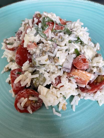 A close-up of a colorful plate of rice salad featuring mixed ingredients such as cherry tomatoes, bell peppers, herbs, and a creamy dressing, served on a turquoise plate.