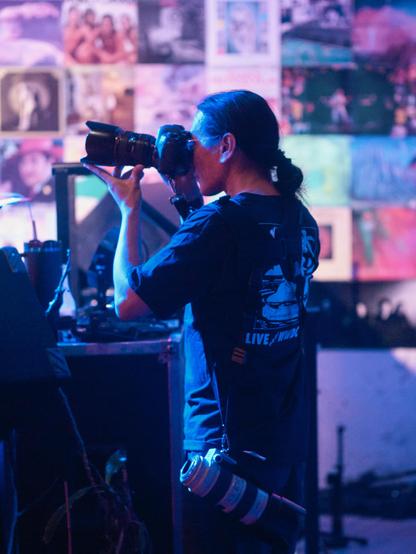 Adam Tow facing left in profile, taking a photo with a camera with a large telephoto lens. He is wearing a vintage LIVE near WWDC t-shirt. Photo by Greg Heo.