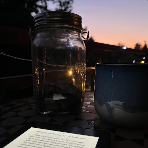 Stillleben in der Sonnenuntergangs-Dämmerung: ein Mosaiktisch mit einer Tasse, einem eBook und einem Glasgefäß (einer Solarlampe). Lichter einer Lichterkette.