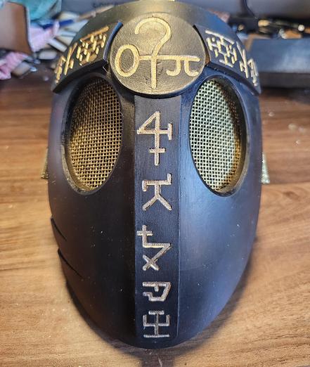 A black and gold 3d printed mask with gold mesh eyes and symbols across its surface 
