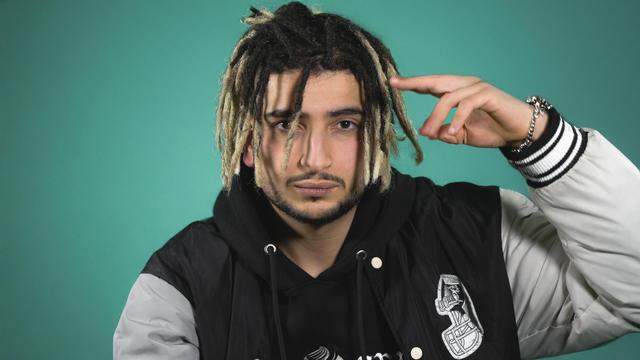 Retrato de primer plano de Buman The General, un hombre joven con rastas teñidas de rubio y negro, chaqueta deportiva y sudadera negra, posando con la mano derecha levantada haciendo su famoso saludo militar de el General sobre un fondo verde turquesa.