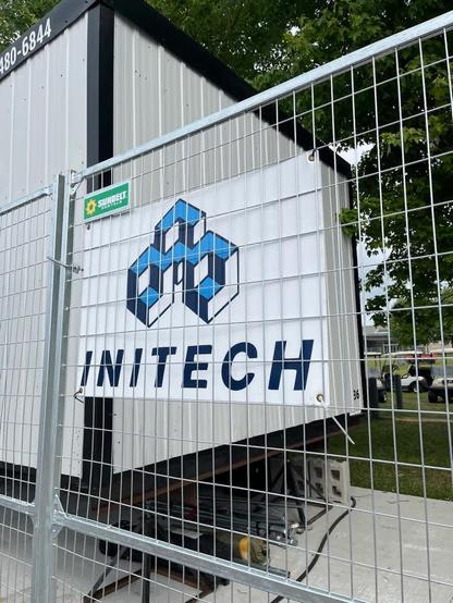 Temporary fencing with a construction trailer behind it and some grass and trees. Attached to the fence is a sign for "INITECH" and a bland corporate logo, which is from the classic 1999 movie Office Space.
