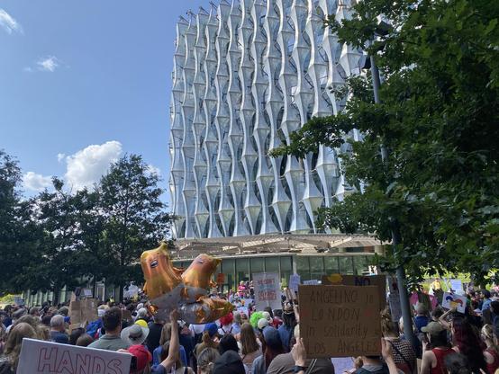A large crowd of people protesting outside the US Embassy in London, which is a large white modern building with diamond shaped elements making up its facade. Three Big Baby Trump balloons are at the center. 