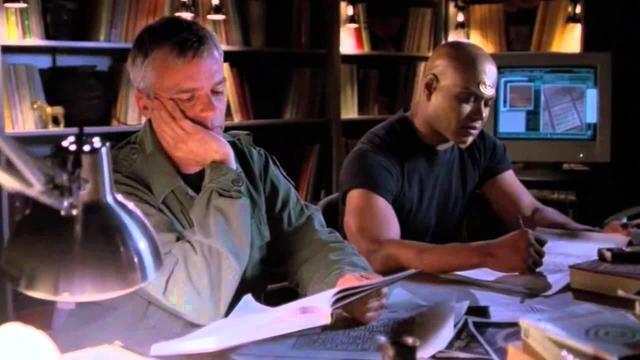 Colonel O’Neill and Teal’c look bored as hell as they study several documents at a table helping Daniel translate alien text