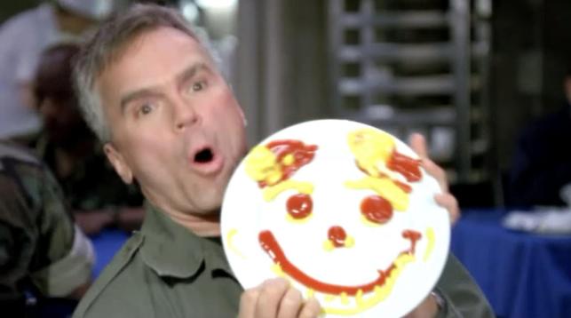 Colonel O’Neill making a crazy face and drawing a smiley face on a plate with ketchup and mustard