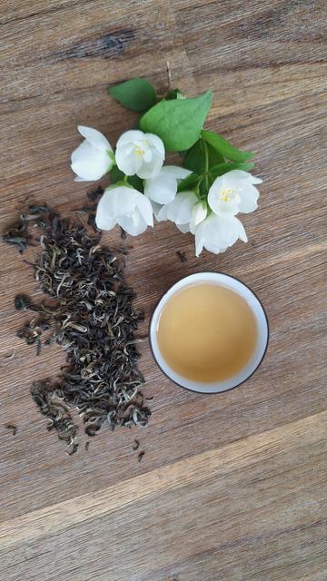 Wooden surface, view from the top. To the left, dry green tea leaves, to the right a brown cup, white inside, filled with green-yellowish tea, towards the top a bunch of white jasmine flowers