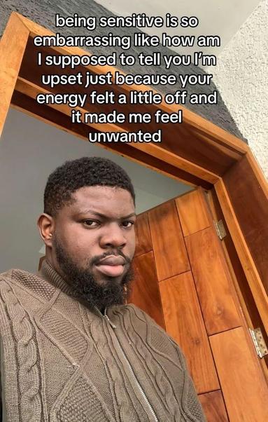 The image features a man standing in a doorway. He has short, curly black hair and a full beard. He is wearing a brown cable-knit sweater with a zipper. The background shows a wooden door and a white wall. The man is looking directly at the camera with a neutral expression. Overlaid on the image is text that reads: "being sensitive is so embarrassing like how am I supposed to tell you I'm upset just because your energy felt a little off and it made me feel unwanted."
