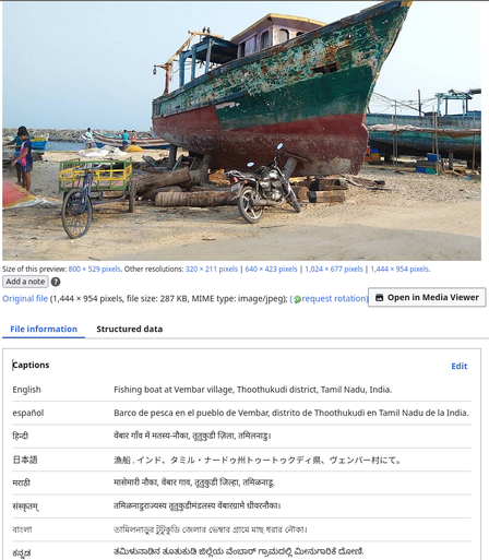 A large, weathered fishing boat on a sandy beach, with its hull painted in green and red, and showing significant signs of wear and rust. The boat is supported by wooden logs and is not currently in the water. In the foreground, a bicycle with a yellow and green cart is parked next to a motorcycle, both resting on the sand. The background features other boats and a few people because it is a fishing village setting. The sky is clear and the overall atmosphere is one of a working coastal area. The image is accompanied by a caption in English: "Fishing boat at Vembar village, Thoothukudi district, Tamil Nadu, India." Additional captions in various languages provide translations of the main caption.
