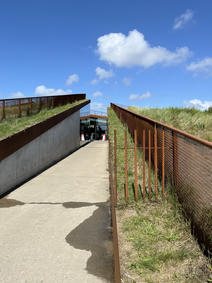 Ein Weg aus Beton in ein Gebäude, welches sich aus der Landschaft mit begrünten Dächern heraushebt. Die Dächer sind begehbar und haben dadurch umlaufende Absturzsicherungen aus Cortenstahl.