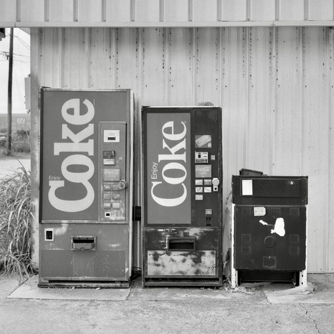 A black and white film photo. Two old soda vending machines sit side-by-side with an old, discarded stove in Taylor, Texas. Sunday, June 15, 2025.
