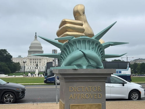 PHOTO: Statue  on the mall with the US Capitol building in the background. The statue shows a golden fist, thumbs up, pounding the cracked head of Lady Liberty, with a broken crown. Beneath, on a pedestal the words "Dictator Approved".