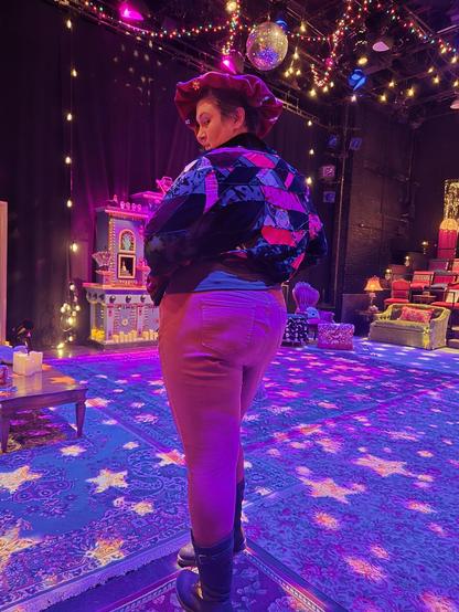 A figure stands in a theater dressed like a seance parlor meets disco club. They are wearing kneehigh boots, red pants, and a high cropped coat that invokes a Shakespearean doublet type costume. On their head is one of those poofy mushroom cap type hats, so the effect is "Shakepeare got lost in a Hot Topic"