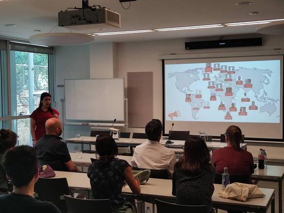 Una mujer está dando una presentación en una sala de conferencias. En la pantalla hay un mapa mundial con fotos de personas y líneas que las conectan, sugiriendo una red global. La presentadora está de pie frente a una pizarra blanca y un grupo de personas sentadas en mesas escuchando atentamente.  Hay un proyector en el techo de la sala.