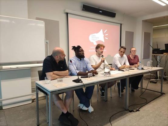 Una mesa larga con cinco personas sentadas en ella, participando en un panel de discusión. Detrás de ellos hay una pantalla de proyección con el título "Data Workers Inquiry" y un icono de megáfono.