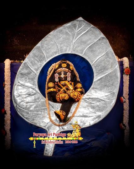 Idol of Hindu deity Krishna in black posing as an infant, right leg folded and tucked behind left knee, relaxing back into a large silver Banyan leaf as if being cradled as someone sings a lullaby. The center of the leaf, the area Krishna leans back onto, is blue. Krishna wears gold shoes, many gold ornaments, and a thin string of brown flowers all around him.

Blue cloth background. Thick strings of white flowers hang down on both ends of the background.