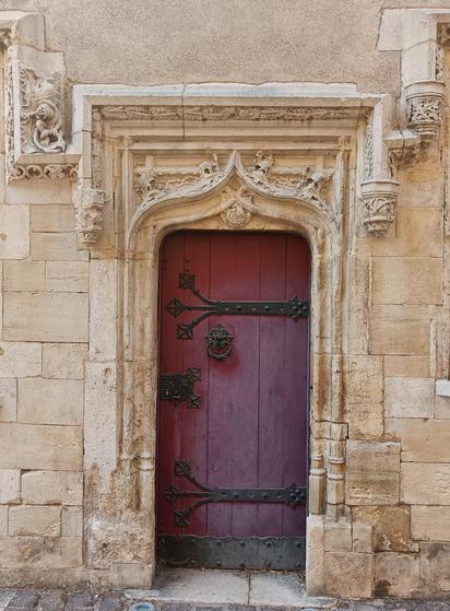 Red wooden door with black castiron hinges, knocker and lock in decorative stone frame. Late medieval style with arch, plant, animal and small figures carved around and small pilasters 